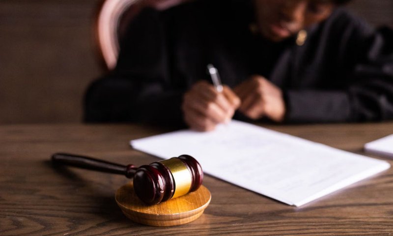 Wooden judge’s gavel on a desk with a judge in a black robe writing in the background